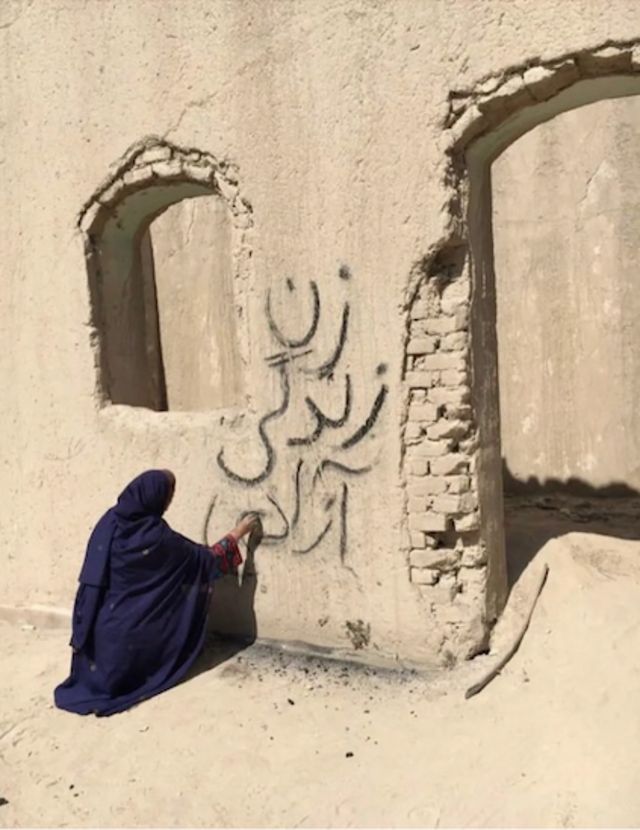 زنی با پوشش چادر روی دیوار کاه‌گلی می‌نویسد: زن، زندگی، آزادی