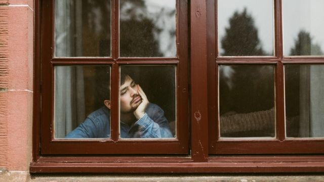 Homem olha pela janela com cara triste
