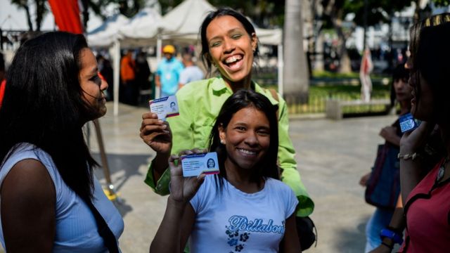 Dos mujeres sonríen a la cámara mientras muestran su carnet de la patria.