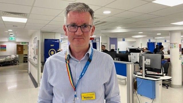 Matthew Trainer, the chief executive of Barking, Havering and Redbridge University Hospitals Trust stands in a hospital. He has glasses on and is wearing a light blue and white striped shirt, a rainbow lanyard and a yellow name badge. In the background there is the hospital reception area and some people sitting at their PCs.