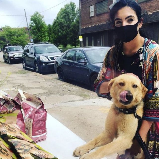 Color photograph shows a woman in a mask next to a mixed-breed, caramel-colored dog