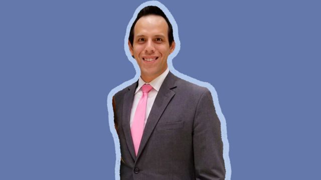 Brett Chapman em pé, de terno e gravata, e fundo azul claro