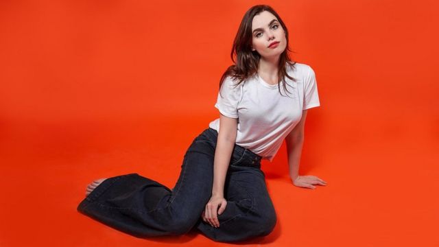 Charli posing in front of a burnt orange background, wearing some worn-look jeans and a white cotton t-shirt