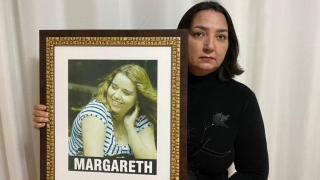 Ruth segura quadro com foto da irmã, Margareth, com olhar sério dentro da sala de casa