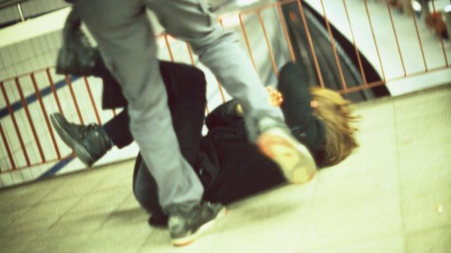 Foto colorida mostra as pernas de uma pessoa que está chutando outra pessoa caída no chão