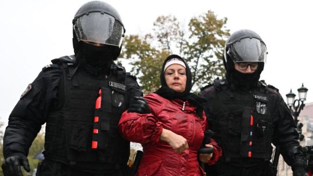 Uma mulher branca de roupa de frio vermelha e lenço preto no cabelo é levada por homens com uniformes e capacetes pretos