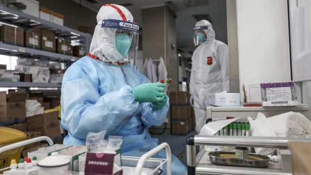 Um profissional de saúde sentado com tubos de amostras é observado por colega ao fundo dentro de laboratório
