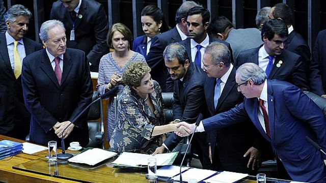 Dilma e Eduardo Cunha apertando as mãos diante da mesa da Câmara dos Deputados, rodeados por outras pessoas