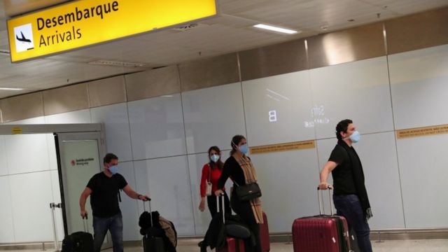 Quatro passageiros com máscaras arrastam suas malas em área de desembarque de aeroporto