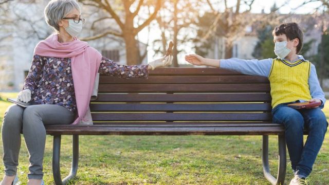 Mujer mayor y niño con mascarillas sentados en un banco de parque.