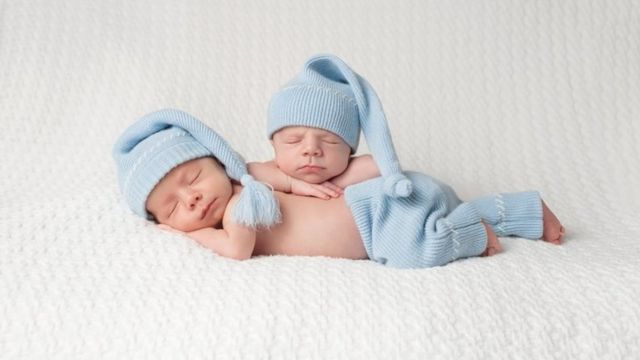 One month old baby fraternal twin boys wearing light blue stocking caps and sleeping on a white blanket
