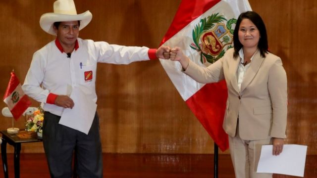 Pedro Castillo e Keiko Fujimori se cumprimentam com mão, dando sorrisos contidos, em frente a bandeira do Peru