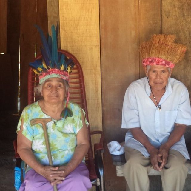 Os idosos Känä́tsɨ e Híwa, últimos falantes da língua warázu, sentados em frente à sua casa em RO, com a acessórios de seu povo indígena na cabeça