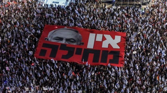 Foto aérea mostra centenas de manifestantes com bandeiras de Israel e um grande pedaço de tecido com o rosto de Netanyahu