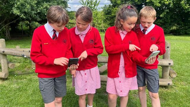 School children in red jumpers and school uniform stood in a park looking at mobile phones playing the game