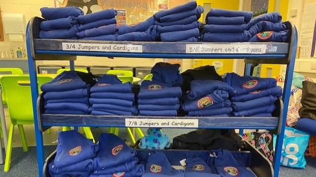 A rack of shelving containing school uniform jumpers. They are labelled for the different school years.