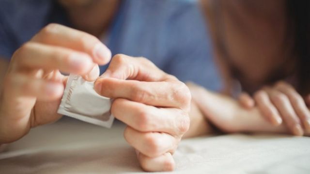 Mão masculina abrindo embalagem de camisinha, com mulher deitada ao lado