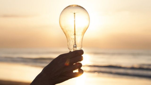 Mão segurando lâmpada diante do sol em uma praia