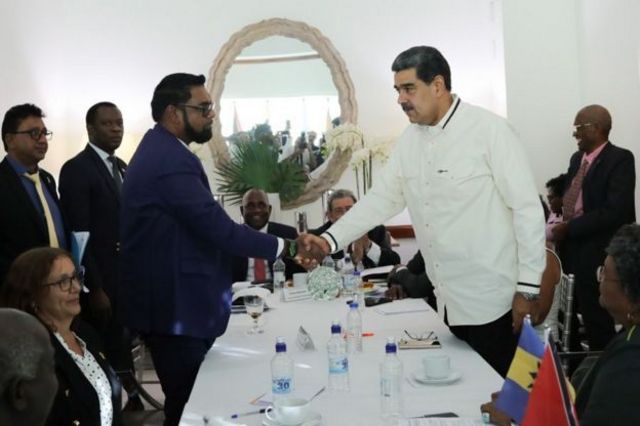 Presidentes da Guiana e Venezuela apertando as mãos, observados por outras pessoas em sala de reunião 