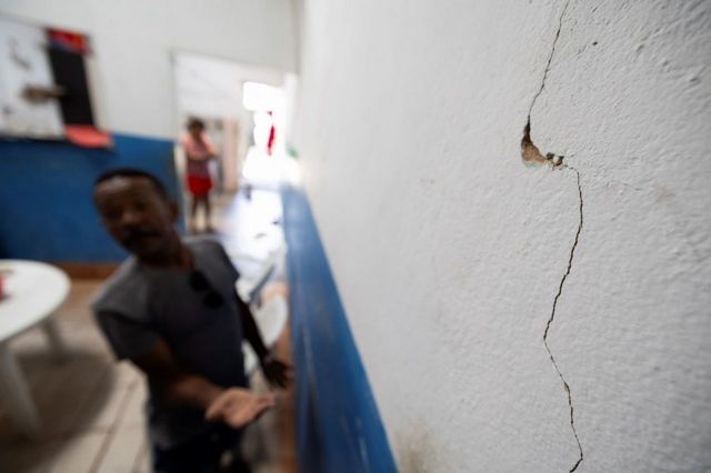 Homem gesticula perto de parede rachada