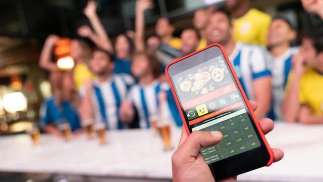 Celular com aplicativo de apostas, e vários torcedores atrás assistindo a jogo em bar