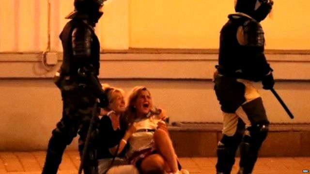 Two women on the floor, screaming, while two policemen with batons stand next to them