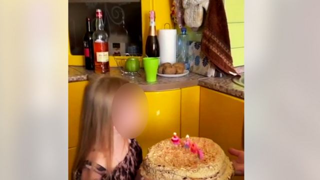 Girl (face blurred) blows out candles on a birthday cake in yellow kitchen