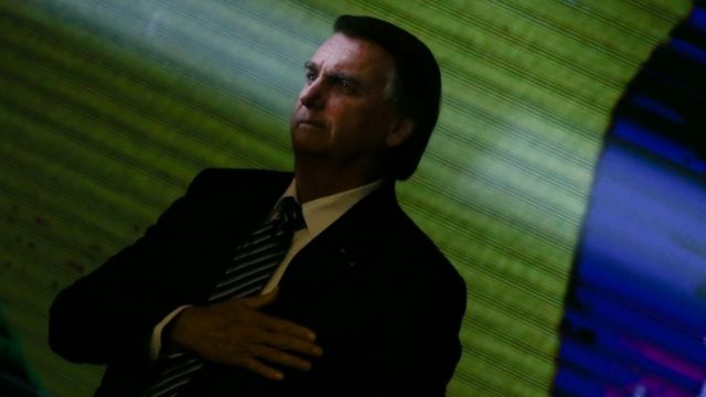 Bolsonaro com mão no peito em frente a telão com bandeira do Brasil