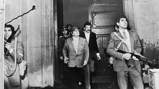 Foto em preto e branco mostra vários homens armados, entre eles Allende, com fuzil na mão e capacete, em pleno ataque ao palácio La Moneda