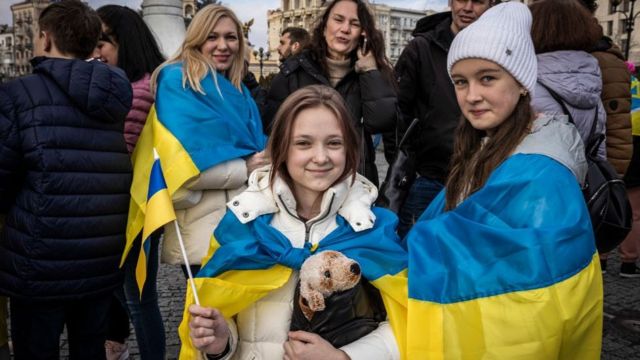 Wanita Ukraina mengibarkan bendera dan tersenyum di depan kamera.