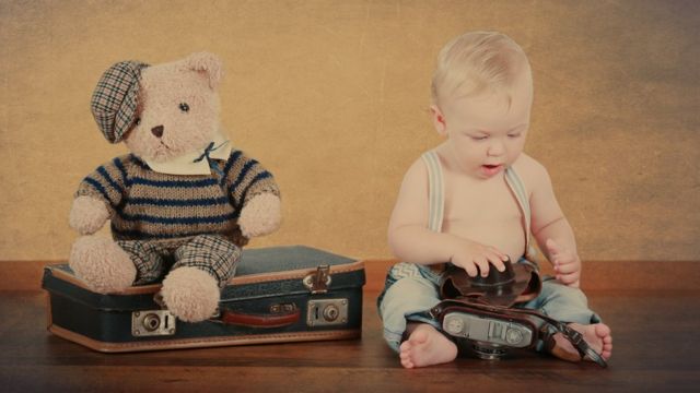 Un bebé con una cámara antigua, un osito de peluche y una maleta