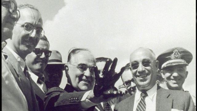 Getúlio Vargas sorri ao mostrar mão suja de petróleo, rodeado por vários homens engravatados