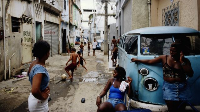 Mulheres conversam apoiadas em uma caminhonete enquanto crianças brincam ao fundo na favela do Jacarezinho, nos anos 1990