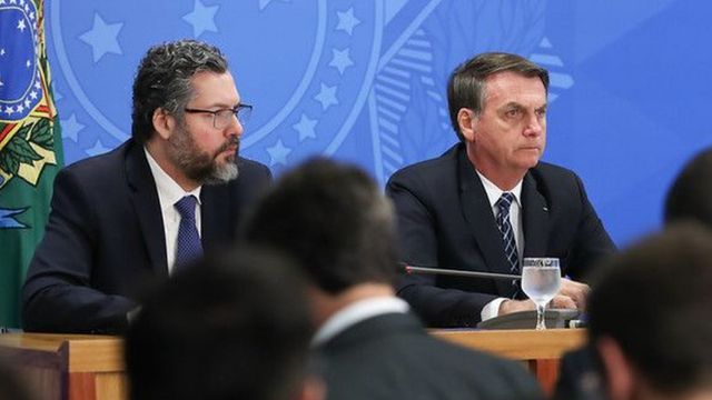Ernesto Araújo e Bolsonaro lado a lado em mesa de evento em ambiente interno