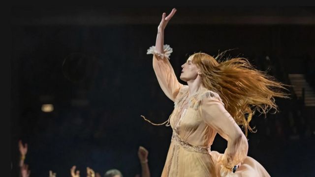 Fotografia mostra Florence Welch, uma mulher branca e ruiva de cabelos muito longos com um vestido rosa vintage dançando sobre um palco, com o fundo negro