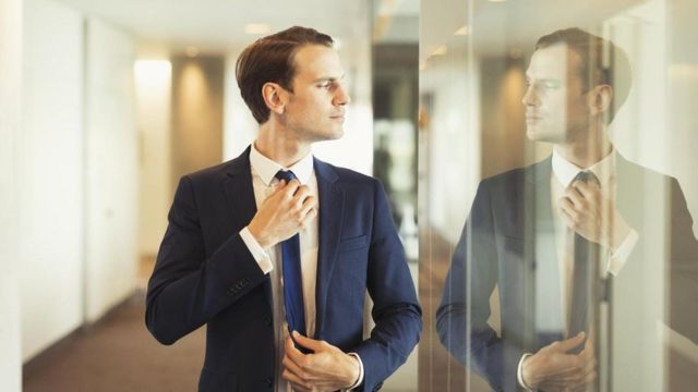 Una persona ajustándose la corbata viéndose en un panel de vidrio de una oficina