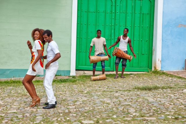 Pareja bailando ta mientras dos hombres, al fondo, tocan tambores. 