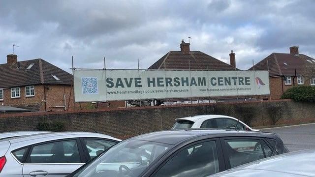 A car park with a banner taped to a fence which reads "Save Hersham Centre"