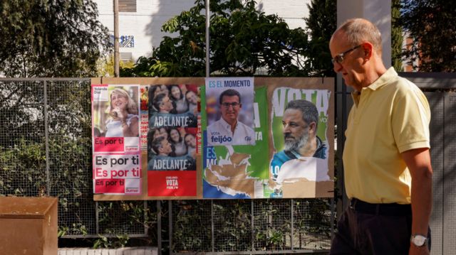 Un hombre camina por una calle junto a una cerca donde hay carteles electorales de varios partidos en España.