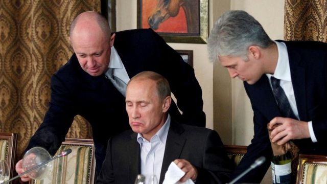 Yevgeny Prigozhin em pé, servindo prato a Putin, que está sentado e faz cara de impressionado