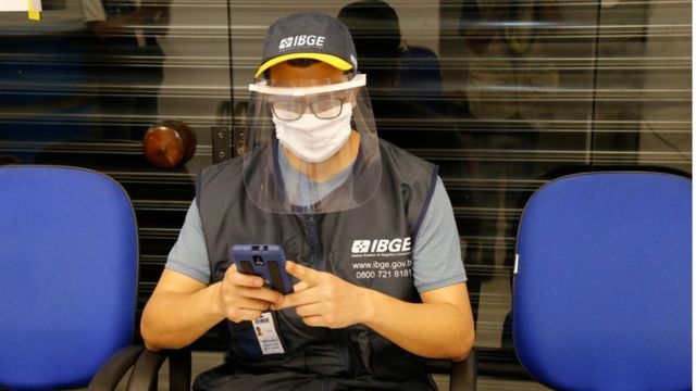 Agente do censo usando boné e colete do IBGE, além de máscara de proteção e face shield contra o coronavírus