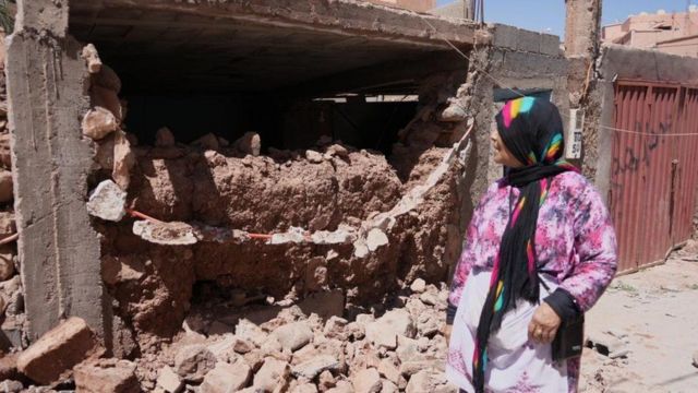 Una mujer se para frente a una casa destruida.