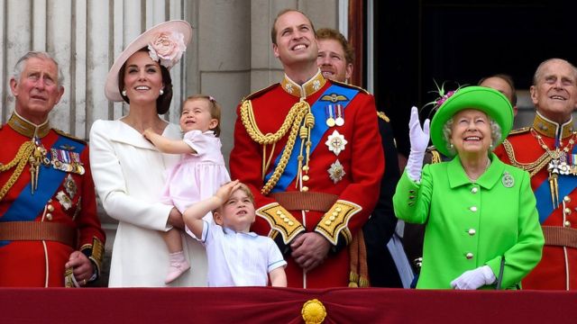 King Charles III, Duchess of Cambridge, Princess Charlotte, Prince George, Prince William, Duke of Cambridge, Prince Harry, Queen Elizabeth II and Prince Philip stand on a balcony.