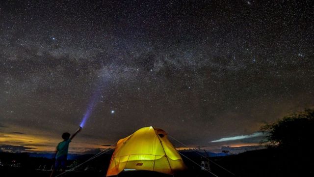 Menino em acampamento à noite com lanterna apontando para céu estrelado