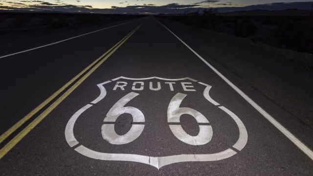 Foto colorida mostra uma sinalização no asfalto de uma estrada que diz "route 66" sob um céu de amanhecer