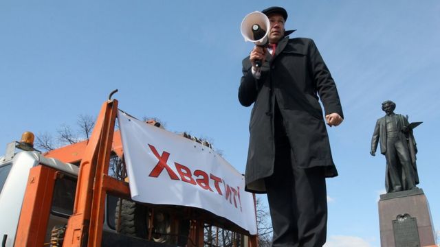 Адвокат Игорь Трунов стоит на импровизированной трибуне с мегафоном в руке на фоне напдиси "Хватит" и памятника Алексаднру Пушкину