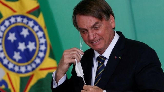 Bolsonaro rindo e tirando a máscara, em frente a bandeira do Brasil