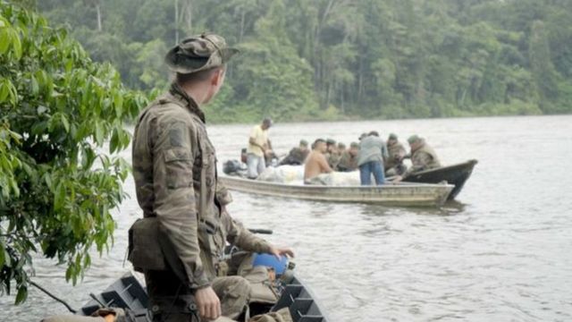 Militar observa homens sendo revistados em barco no meio da floresta