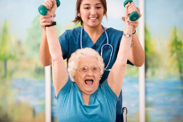 Idosa branca levantando os braços com ajuda de uma jovem com jaleco
centenários
