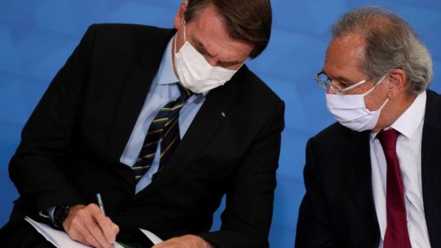 Bolsonaro assinada papel, observado por Paulo Guedes, ambos sentados com máscara em evento